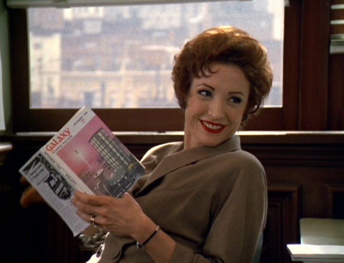 Kay Eaton (played by Nana Visitor) is in a brownish-gray outfit, sitting down while smiling and holding a copy of the sci-fi magazine Galaxy.