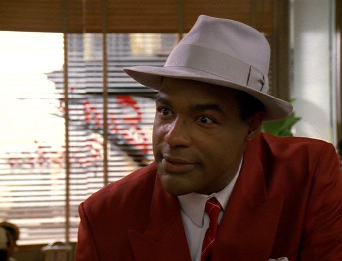 Willie Hawkins (played by Michael Dorn) is in a red suit and gray hat and has an expression of real interest on his face.