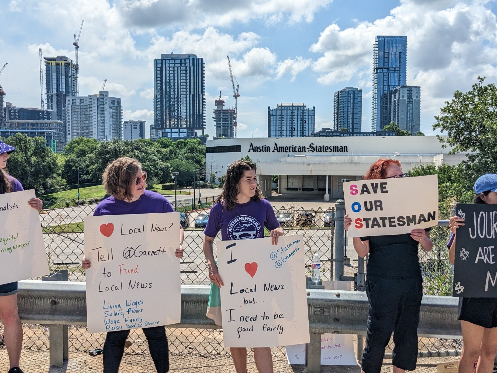 Protesters stand on the Congress Avenue bridge with the Austin skyline behind them, holding signs with messages about Gannett and the Austin American-Statesman, such as "Save Our Statesman" and "I (heart) local news but I need to be paid fairly".