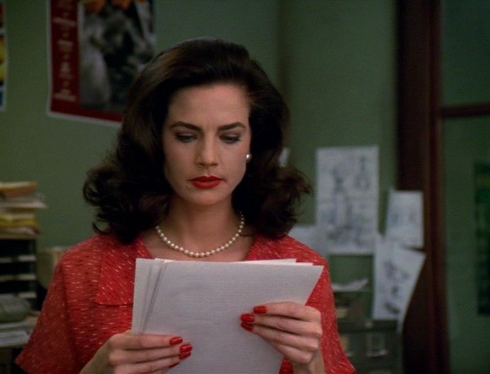 Darlene Kursky (played by Terry Farrell) is wearing a red outfit with a pearl necklace and she’s reading from a stack of pages that she’s holding.